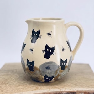 Op de afbeelding: Een crèmekleurige keramische kan met een herhalend patroon van zwarte kattengezichten met grote ogen en kleine zwarte bijen. De onderkant van de kan heeft gevlekte grijze en bruine cirkelvormige vlekken, inclusief een gebogen handvat en een tuit.
