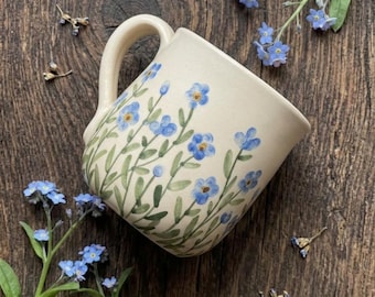 May include: A cream-colored ceramic mug featuring a hand-painted pattern of small blue forget-me-not flowers with green stems and leaves. The mug has a curved handle and a smooth finish, decorated with multiple floral clusters around the exterior.