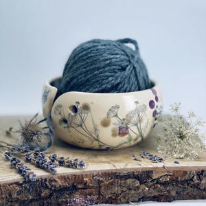 Könnte beinhalten: Eine cremefarbene Keramik-Garnschale mit einem botanischen Aufdruck von Wildblumen und einer spiralförmigen Öffnung. Ein großes Knäuel dunkelblaues Garn liegt in der Schale, wobei ein einzelner Faden durch die seitliche Öffnung geführt wird.