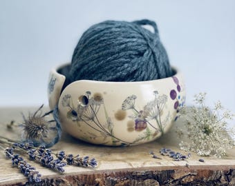 May include: A cream-colored ceramic yarn bowl featuring a botanical print of wildflowers and a spiral cutout for yarn. A large ball of dark blue yarn sits inside the bowl, with a single strand threaded through the side opening of the vessel.