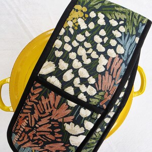 May include: Floral oven mitt with a black border. The mitt features a dark blue background with a pattern of white, yellow, and rust-colored flowers and green leaves. The text "Mo me" is visible on the side.