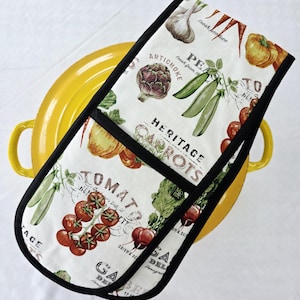 May include: A white oven mitt with a black trim, featuring a vintage-style print of various vegetables like tomatoes, carrots, and artichokes. The text includes "Heritage Carrots" and "Fresh Produce". The mitt is placed over a yellow pot.