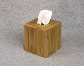 May include: A wooden tissue box holder with a light brown wood grain finish. A white tissue is partially pulled out of the box.