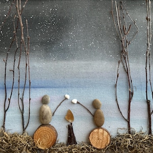 May include: A winter scene with two stone figures holding marshmallows over a small fire. The background is a blue and gray sky with white snow falling. The figures are sitting on wooden logs with moss around them. The scene is made with natural materials.