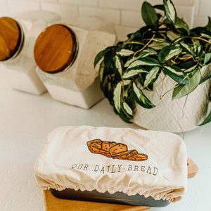 May include: A loaf of bread in a dark gray bread pan, covered by a cream-colored cloth with a bread illustration and the text "OUR DAILY BREAD". The pan sits on a wooden cutting board. A plant and canisters are in the background.