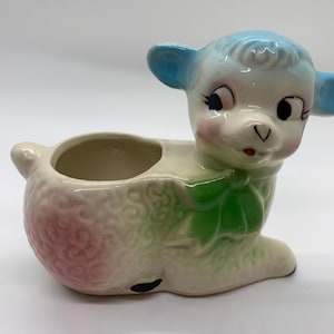 May include: A ceramic planter shaped like a lamb with a glossy finish. The lamb has light blue ears, a white face with pink cheeks, and a textured body featuring patches of soft pink and green. A green bow is painted around the neck area.