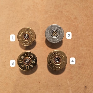 May include: Four shotgun shell casings, each with distinct markings. Two are brass-colored, one is silver, and one is brass with the text "FIOCCHI ITALY 12". The casings are arranged on a wooden surface.