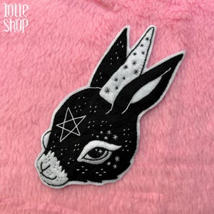 May include: A black and white felt rabbit applique featuring a pentagram and star details. The rabbit has a detailed eye and is set against a pink background. The text "LOVE SHOP" is in the upper left corner.