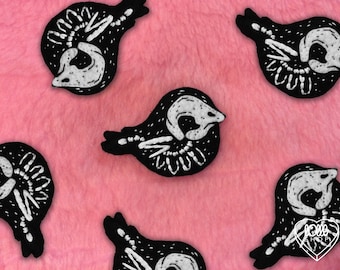 Puede incluir: Múltiples parches bordados negros con forma de pájaro y detalles esqueléticos blancos. Cada parche presenta una silueta de pájaro estilizada con hilo blanco que representa el cráneo, las alas y los huesos de la cola sobre tela negra.