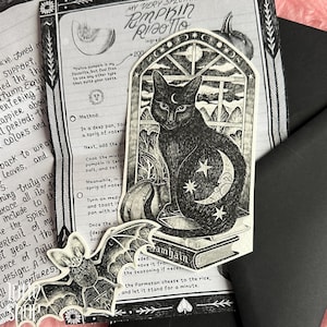 Op de afbeelding: Een zwart-witte sticker met een kat die op een boek met het label Samhain voor een raam zit, met een maansikkel en sterren op de vacht. Een aparte vleermuissticker met de tekst Creature is linksonder op de afbeelding geplaatst.