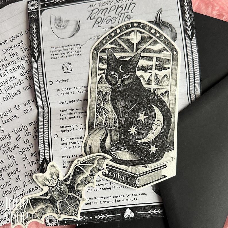 Op de afbeelding: Een zwart-witte sticker met een kat die op een boek met het label Samhain voor een raam zit, met een maansikkel en sterren op de vacht. Een aparte vleermuissticker met de tekst Creature is linksonder op de afbeelding geplaatst.