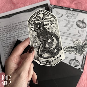 Op de afbeelding: Een gestanste sticker met een zwarte kat die op een stapel boeken naast een pompoen zit, omlijst door een boogvenster. De kat heeft een maansikkel op het voorhoofd en een maan en sterren op het lichaam. Het woord Samhain staat onderaan gedrukt.