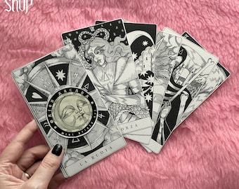 Op de afbeelding: Een set tarotkaarten met zwart-wit illustraties. De kaarten liggen uitgespreid op een roze pluche oppervlak. De kaarten omvatten het Rad van Fortuin, de Toren, de Dood en de Ster.