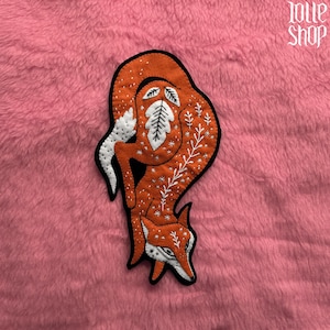 Può includere: Una toppa a forma di volpe arancione con dettagli bianchi e neri. La volpe è decorata con disegni di foglie e rami bianchi. La toppa è su una superficie rosa e sfocata. Il testo "lolle shop" si trova nell'angolo in alto a destra.