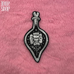 Può includere: Toppa ricamata in bianco e nero a forma di bottiglia di pozione. La toppa presenta un disegno floreale con un teschio al centro. La toppa è su uno sfondo rosa e sfocato. Il testo "lolle shop" è nell'angolo in alto a sinistra.