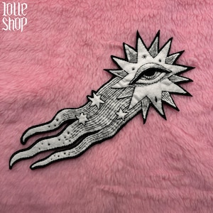 以下が含まれることがあります： 中心に目がついた流れ星の黒いアウトラインが描かれた白い刺繍パッチ。星は下から涙の形をしています。パッチはピンクのふわふわした背景の上にあります。