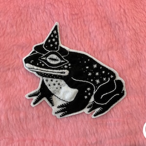 May include: Black embroidered patch of a frog wearing a pointed hat with stars on it. The frog is outlined in white and has a white eye. The background is pink.