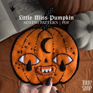 Puede incluir: Un adorno de fieltro naranja con forma de calabaza, detalles cosidos en negro, una luna creciente negra en la frente, ojos blancos y naranjas con lágrimas negras y una boca ancha con dientes blancos. El texto dice Little Miss Pumpkin SEWING PATTERN | PDF.