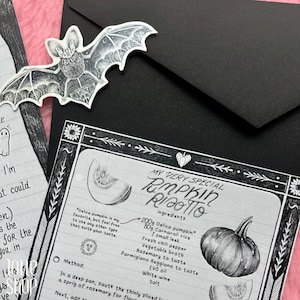 Op de afbeelding: Een zwart-wit receptenkaart voor pompoenrisotto met handgetekende illustraties van een pompoen, een vleermuisvormige sticker met de tekst Autumn Creature en een zwarte envelop op een roze gestructureerde ondergrond.