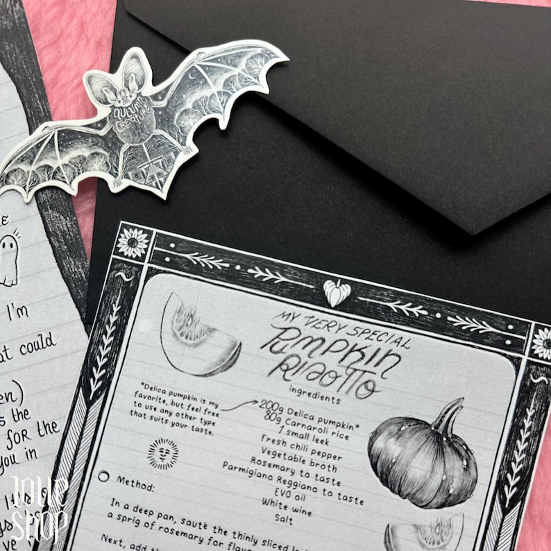 Op de afbeelding: Een zwart-wit receptenkaart voor pompoenrisotto met handgetekende illustraties van een pompoen, een vleermuisvormige sticker met de tekst Autumn Creature en een zwarte envelop op een roze gestructureerde ondergrond.