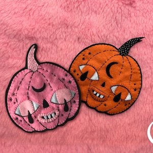 Può includere: Due decorazioni in feltro a forma di zucca, una rosa e una arancione, con dettagli cuciti, mezzelune nere sulla fronte, occhi triangolari bianchi con lacrime nere e piccole bocche con denti bianchi. Ogni zucca ha un picciolo nero.