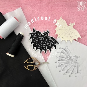 Può includere: Un progetto artigianale con una toppa a forma di pipistrello nera con stelle bianche, una toppa color crema con stelle nere e un'illustrazione di pipistrello su carta. Include rocchetti di filo bianco e nero, forbici dorate e tessuto nero.