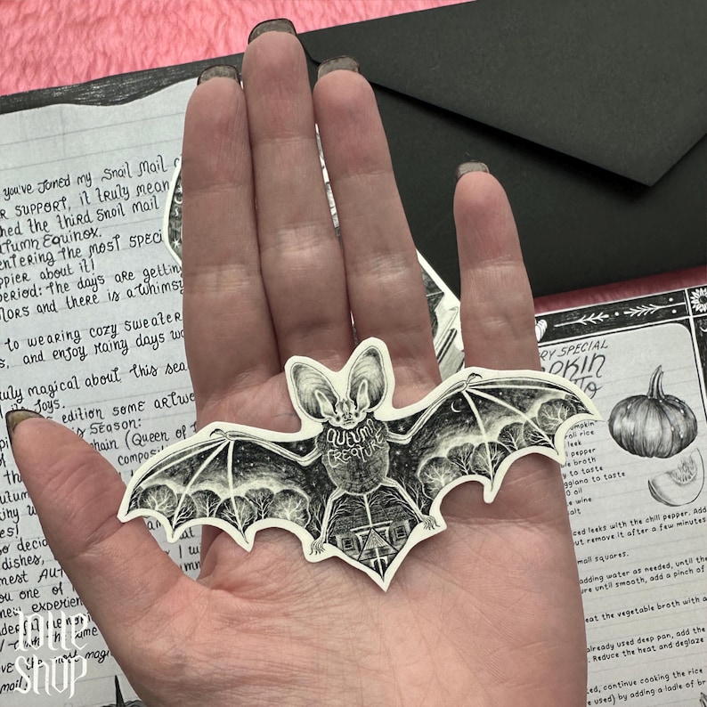 Op de afbeelding: Een gestanste sticker in de vorm van een vleermuis met zwart-witte inktillustraties. Het lichaam van de vleermuis bevat de tekst 'AUTUMN CREATURE' en een gedetailleerde sc&egrave;ne van een huis en bomen onder een sterrenhemel met een maansikkel.