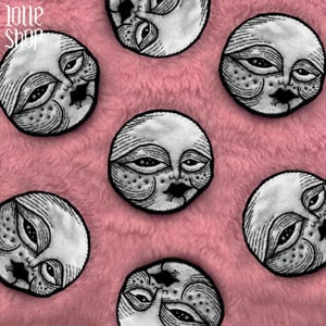 May include: Embroidered moon face patches with detailed black linework and borders. The patches are white and black, set against a pink, fluffy background. The text "Jone Shop" is in the upper left corner. These patches are circular in shape.