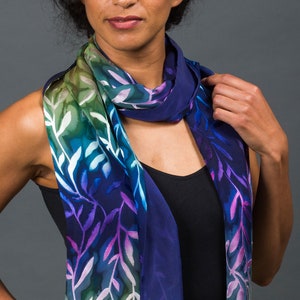 May include: A silk scarf with a leafy pattern in shades of green, blue, purple, and white. The scarf is draped around the neck of a person wearing a black top.