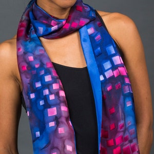 Op de afbeelding: Een blauwe en roze zijden sjaal met een geometrisch patroon. De sjaal is om de nek van een persoon gebonden die een zwart tanktop draagt.