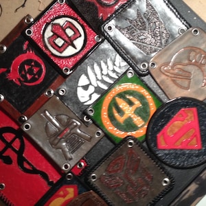 Könnte beinhalten: Eine Sammlung von Lederpatches mit verschiedenen Designs, darunter ein Superman-Logo, ein Farn, ein Dreizack und ein Schädel. Die Patches sind auf einem schwarzen Hintergrund zusammengenäht.