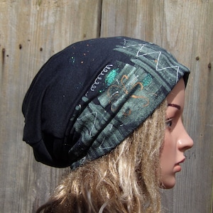 May include: A black beanie hat with a patterned design in shades of green, white, and orange. The hat has a folded brim and is made of a soft, comfortable material. The design includes abstract shapes and lines.