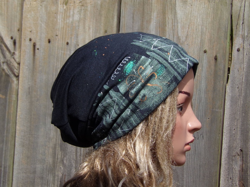 May include: A black beanie hat with a patterned design in shades of green, white, and orange. The hat has a folded brim and is made of a soft, comfortable material. The design includes abstract shapes and lines.