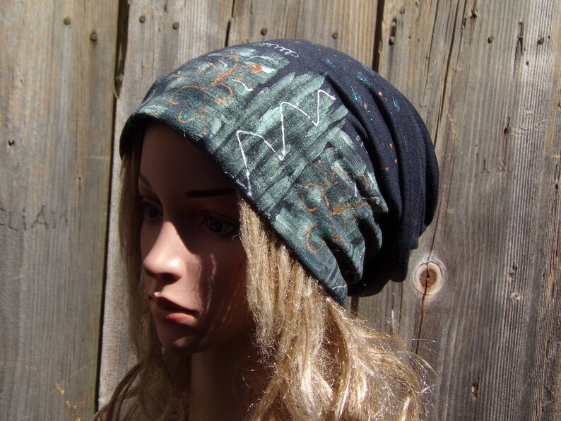 May include: A dark blue beanie hat with a green and white abstract design. The hat has a folded brim and is made of a soft, stretchy fabric. The design features geometric shapes and orange accents.
