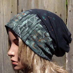 May include: A dark gray beanie with a painted design. The design features green brushstrokes, white zigzags, and orange accents. The beanie is made of a soft material and is suitable for casual wear.