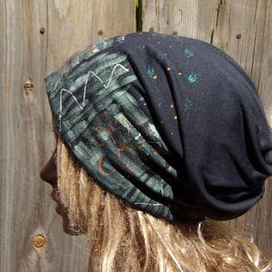 May include: A dark gray beanie hat with a unique design. The hat features a section with a green and black pattern with white and orange accents. The other section of the hat has green and orange paint splatters.