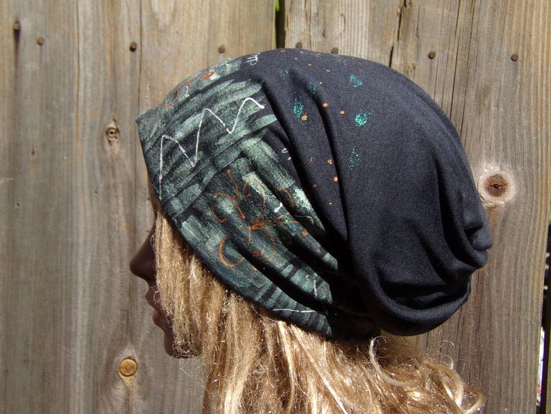 May include: A dark gray beanie hat with a unique design. The hat features a section with a green and black pattern with white and orange accents. The other section of the hat has green and orange paint splatters.