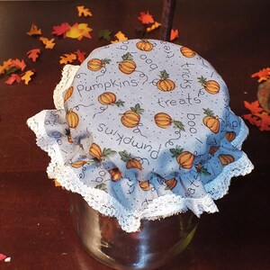 May include: A glass jar covered with a light blue fabric lid featuring orange pumpkins and the words "Pumpkins treats boo". The lid is trimmed with white lace. Small orange pumpkin decorations and fall leaves are scattered around.