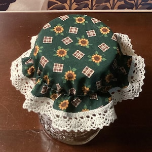 May include: A glass jar covered with a green fabric lid featuring a sunflower pattern and a white lace trim. The background includes a painting of a cow and a sunflower field.