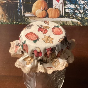 May include: A white fabric jar lid with a fall theme. The lid has a ruffled edge and features a pattern of orange pumpkins, brown leaves, and acorns.
