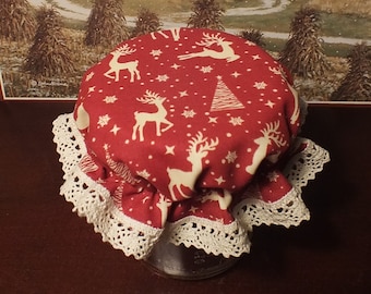 May include: A circular fabric jar cover featuring a red background with a repeating pattern of white reindeer, snowflakes, and stylized trees. The edge of the cover is finished with a decorative white crocheted lace trim that hangs down in soft folds.