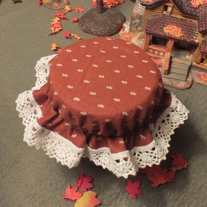 May include: A miniature table setting with a brown tablecloth and white lace trim. The table is surrounded by miniature houses and a small tree with orange leaves. The scene is decorated with small orange and red leaves.