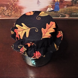 May include: A jar with a black fabric lid featuring a colorful autumn leaf pattern. The background shows a painting of a fall landscape with a river, trees, and figures.