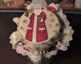 May include: A glass jar covered with a fabric lid featuring a Santa Claus design. The lid is trimmed with white lace. The Santa Claus illustration is on a cream-colored background with holly and berry accents. The jar sits on a dark brown surface.