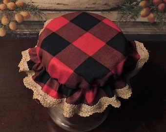May include: A red and black plaid fabric jar lid cover with a cream colored lace trim. The lid cover is sitting on a dark brown surface.