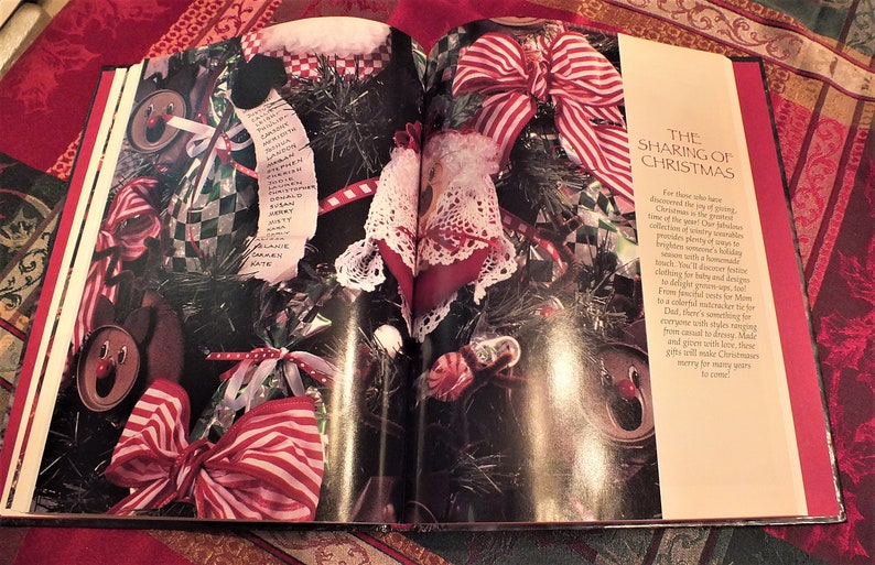 May include: An open book with a red and white striped bow on the cover. The book is titled "The Sharing of Christmas". The text on the page reads "For those who have discovered the joy of giving, Christmas is the greatest time of the year. Our greatest collection of country treasures to brighten your winter holidays. You'll discover a holiday treasure someone's sure to love. You'll discover festive trinkets for baby and grown, from fanciful lamps and signs to a cuddly red cracker for Dad. There's something for everyone with stylish magpies from casual to dress. Made and grown with love, these gifts will make Christmas merry for many years to come!"