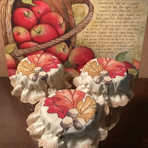 May include: Three glass jars with fabric lids featuring autumn leaf and acorn designs. The lids have a cream-colored base with a lace trim. The background includes a watercolor illustration of apples and a recipe for Emmy's Apple Tart.