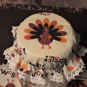 May include: A clear glass jar with a fabric lid featuring a turkey design in shades of brown, orange, and yellow. The lid has a white lace trim and the words "GOBBLE" and "VORIE" printed on it. A crow is perched on a fence in the background.