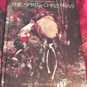 May include: A book titled "The Spirit of Christmas" by Leisure Arts Presents. The cover features a Santa Claus figure made of natural materials, including twigs, leaves, and fabric. The book is a guide to creative holiday ideas.