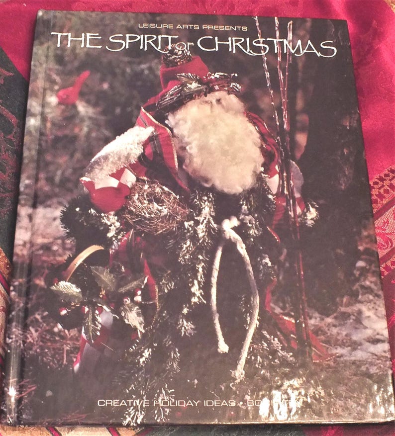 May include: A book titled "The Spirit of Christmas" by Leisure Arts Presents. The cover features a Santa Claus figure made of natural materials, including twigs, leaves, and fabric. The book is a guide to creative holiday ideas.